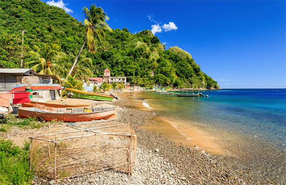 A tropical beach scene with clear blue water, a sandy shore, boats on the sand, and lush greenery on a hillside in the background.