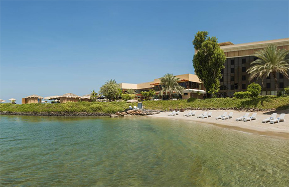 A serene beach with clear water, sandy shore with chairs, low-rise buildings possibly a resort, surrounded by greenery under a blue sky.