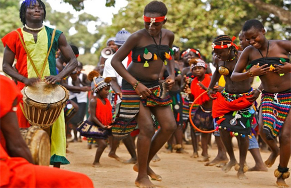 A group of people in colorful traditional African attire dancing and playing drums outdoors.