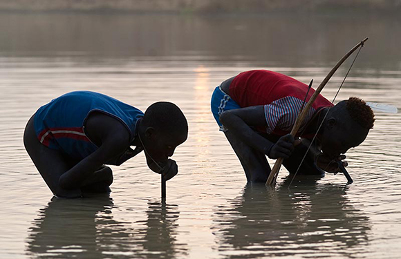 Two people are seen near water, seemingly collecting something in dim light, creating a silhouette effect.