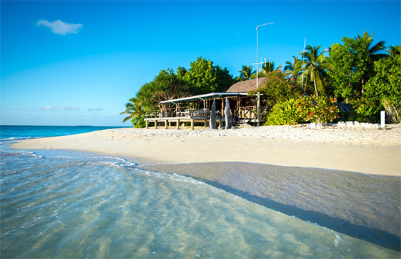 A tropical beach features clear blue water, white sand, a small hut, green palm trees, and a partly cloudy blue sky.