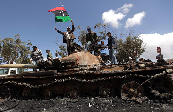 A group of people standing on a destroyed tank with one person waving a flag with a red, black, and green design.