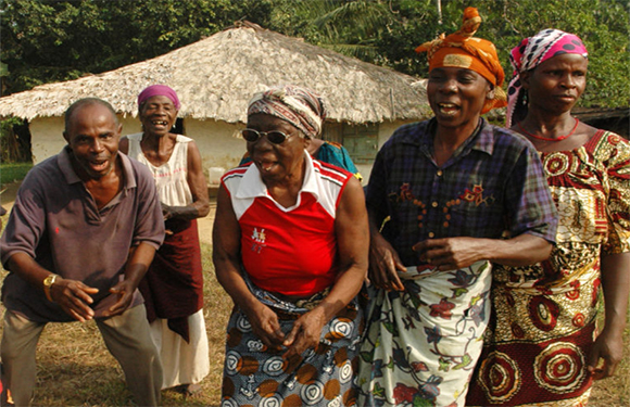 Older African adults pose outdoors in traditional clothing, with a thatched-roof structure behind.