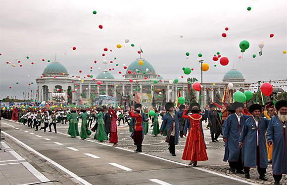 People in traditional attire walk in a parade, releasing green and red balloons, with classical buildings behind.