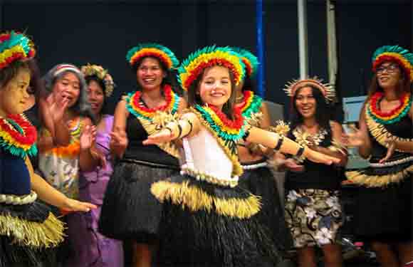 A group in feathered headpieces and grass skirts dance on stage, focusing on a smiling, dancing young girl.