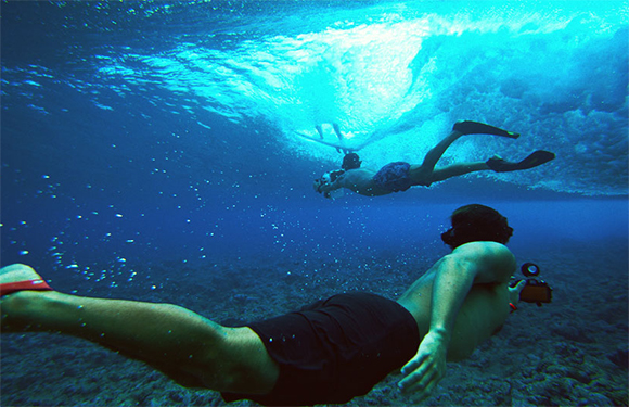 Two people snorkeling underwater, one closer to the camera, with sunlight filtering through the surface of the water.