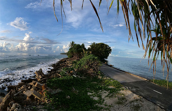 A narrow road leading through a coastal landscape with the ocean on one side and lush greenery on the other, under a partly cloudy sky.