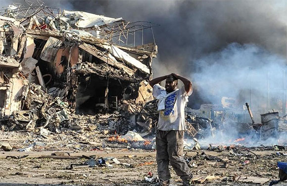 A distressed person stands before a damaged building with debris and smoke, indicating recent disaster.