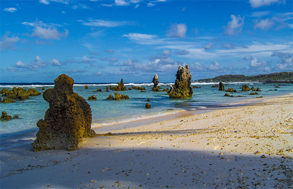 A scenic beach with white sand, rock formations in the shallow water, and a blue sky with clouds.