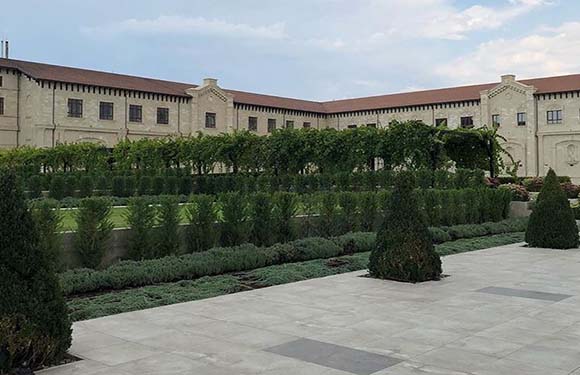 An image of a formal garden with neatly trimmed hedges and topiaries in front of a classic building with arches and tiled roof.