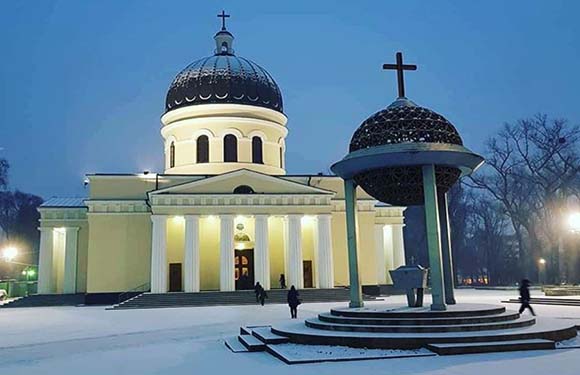 A twilight photo of a domed church in a snowy landscape, with people walking by and a gazebo-like structure nearby.