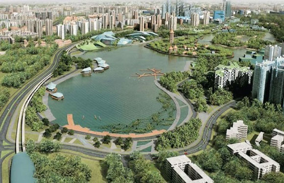 Aerial view of an urban park with a large lake, surrounded by green areas and a variety of buildings, with roads curving around the landscape.