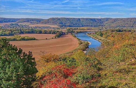 A scenic view of a meandering river flowing through a valley with patches of agricultural fields and surrounded by wooded areas with autumn foliage.