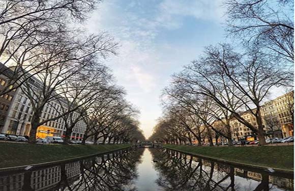 A daytime wide-angle photo of a tree-lined canal in a city, with buildings and tree reflections in the water.