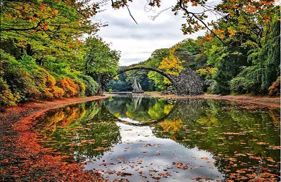 A picturesque scene of a calm lake reflecting a stone arch bridge surrounded by trees with autumn foliage.