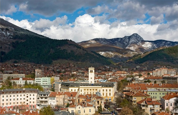 A panoramic view of a town with buildings in the foreground and mountains in the background under a partly cloudy sky.