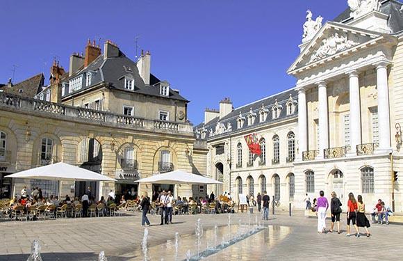 A sunny day at an outdoor plaza with people walking and sitting at cafe tables, with elegant buildings and a small fountain in the foreground.