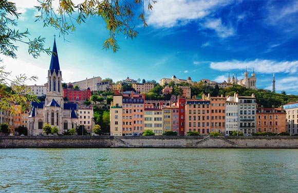 A scenic European cityscape with vibrant buildings by a river, a church spire, a hilltop fortress, under a semi-cloudy sky.