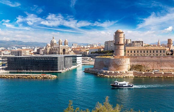 A coastal city with historic structures, a cathedral and tower, overlooks a sea with a sailing boat under a clear, partly cloudy sky.