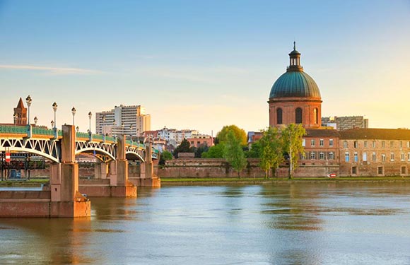 A scenic view of a river with a bridge in the foreground and buildings with a prominent dome structure in the background during sunset or sunrise.