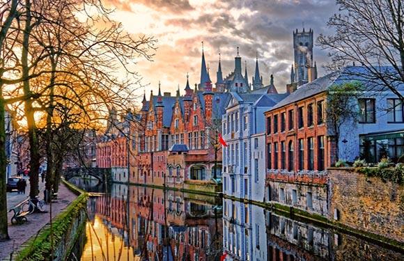 A picturesque view of a European city at sunset with historic buildings reflected in a calm canal, vibrant sky, and trees along the water's edge.