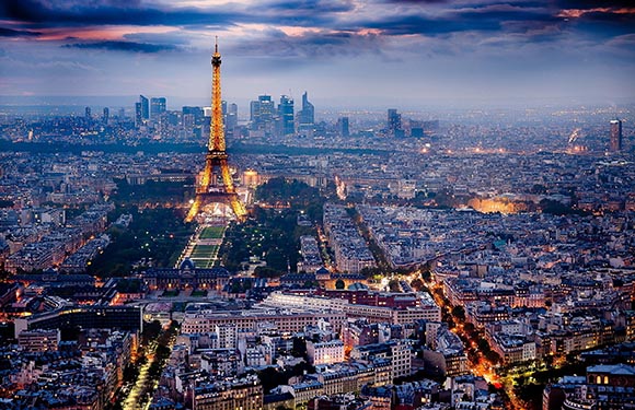 Aerial view of Paris at dusk with the Eiffel Tower illuminated and city lights starting to turn on, with a hazy sky.