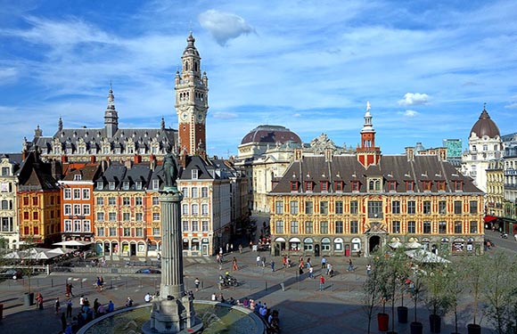 Historical city square with a tower, monument, and distant people under a partly cloudy sky.