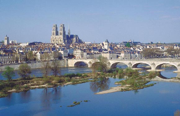 A city panorama featuring a river, stone bridge, historic buildings, a cathedral with two towers, under a clear blue sky.