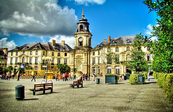 A vibrant town square with people walking, classic European architecture, a prominent clock tower, and a blue sky with fluffy clouds.