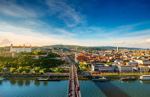 A panoramic view of a city during the daytime with a river running through it, a bridge spanning the river, and a clear blue sky with a few clouds.