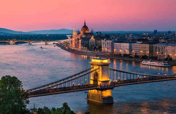 A city at dusk with a river, a prominent bridge, illuminated buildings under a pink and blue sky.