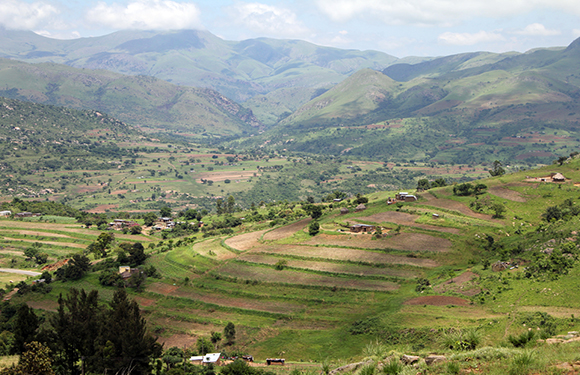 A scenic view of a lush, green valley with terraced fields, dotted with small buildings, surrounded by rolling hills.