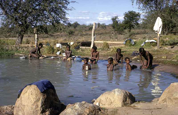 A group of people bathing and washing clothes in a shallow river surrounded by rocks and vegetation under a clear sky.