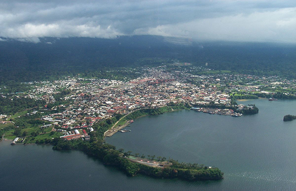 Aerial view of a coastal town with buildings densely packed together, surrounded by water on one side and greenery on the other, under a cloudy sky.