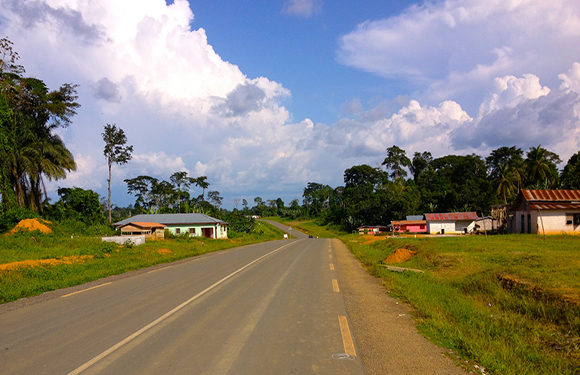 A rural road with a clear sky and scattered clouds above, flanked by greenery and a few small buildings.