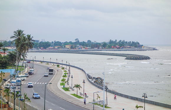 A coastal road curving along the shoreline with a few cars, palm trees lining the side, and buildings in the distance under a cloudy sky.