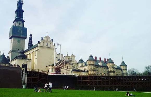 A photo depicts people lounging and strolling on grass before a grand, historic castle under a cloudy sky.