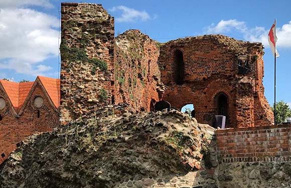 A picture of a decaying red brick castle ruin under a partly cloudy sky, with a flag on the right and greenery at the base.