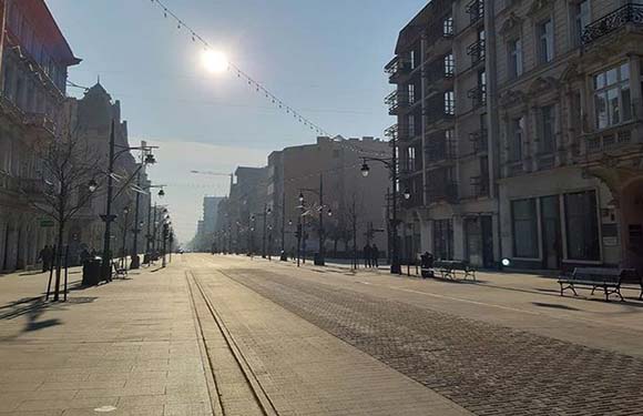 An image of a quiet urban street with buildings on either side, street lamps, benches, and a clear sky.