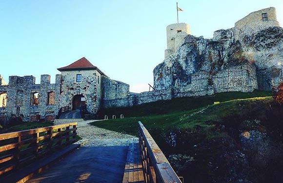 An image of a historic stone castle with a prominent tower, set against a clear sky. A wooden railing leads towards the entrance.