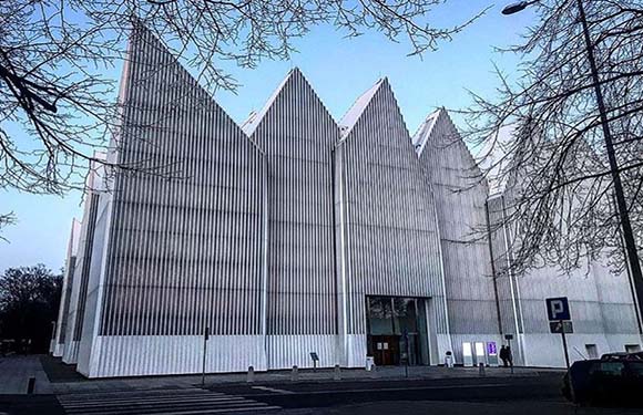 A modern building with a unique pointed roof, vertical facade, partially obscured by trees, with a parking area sign.