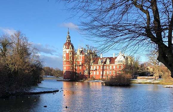 A scenic view of a traditional European castle with spires, mirrored in a moat, framed by trees and a blue sky.