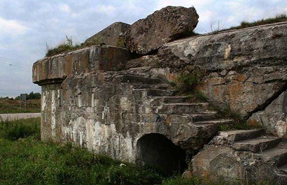 A dilapidated stone structure with a large boulder on top, featuring worn steps on the side and an arched opening at the base.