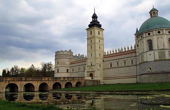 A historical building with a tall tower and a dome, adjacent to a bridge with multiple arches over a body of water, under a cloudy sky.