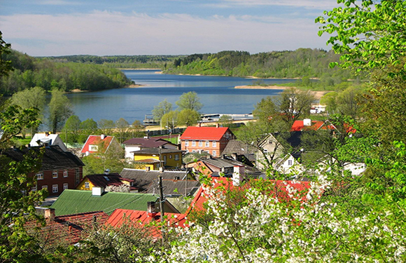 Scenic view of a village with colorful buildings among trees, a lake in the background, and white blossoms upfront.