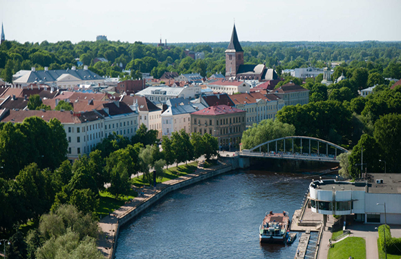 Aerial cityscape with a river, bridge, boat, buildings, a prominent tower, and surrounding greenery.