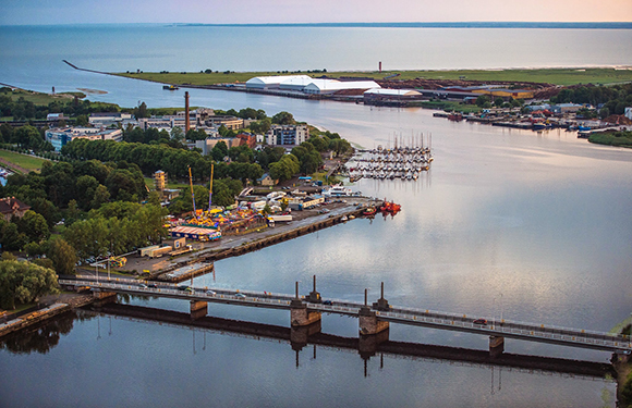 Aerial view of a calm waterfront with a marina full of boats, a bridge in the foreground, and industrial buildings in the distance during twilight.