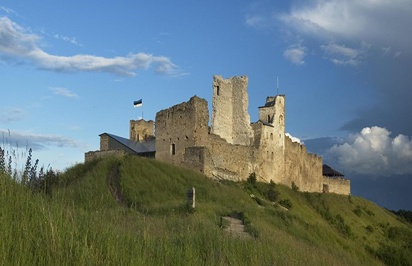 A medieval castle with ruined walls and towers sits atop a grassy hill under a partly cloudy sky.