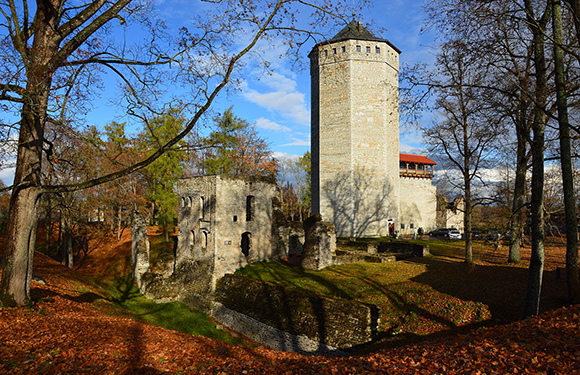 A stone tower and ruins surrounded by bare trees with autumn leaves on the ground under a blue sky with scattered clouds.