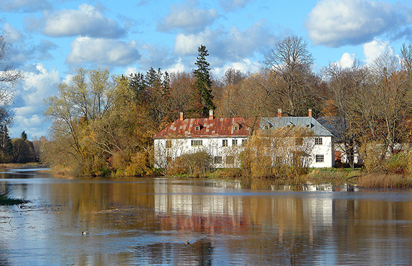 A large white house with a red roof, surrounded by autumn trees, reflects in calm waters under a partly cloudy sky.